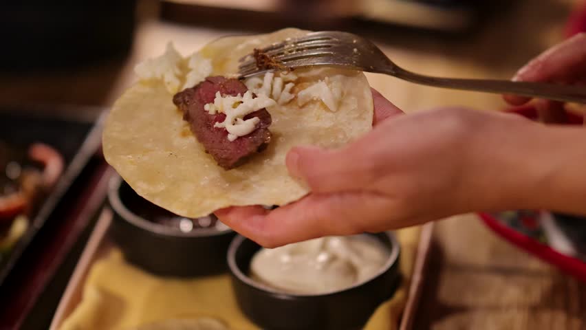 Hands prepare a tortilla with steak, shrimp, and cheese on a hot skillet in a warmly lit restaurant setting, using close-up camera angles