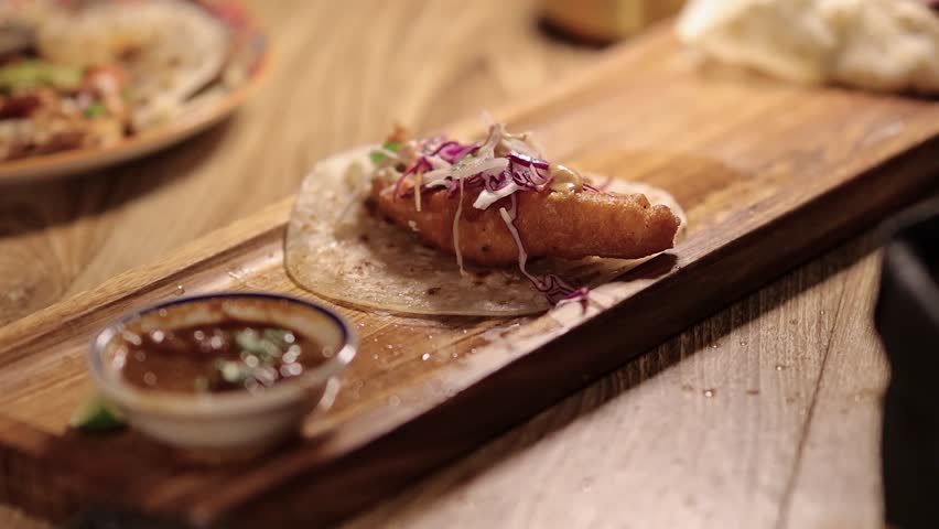 A hand picks up a crispy fish taco topped with slaw from a wooden board in warm, soft lighting. Close-up, shallow depth of field