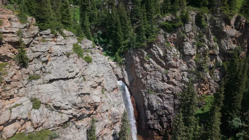 Breathtaking aerial view of a rocky mountain landscape featuring a cascading waterfall surrounded by lush green pine forests under a clear blue sky