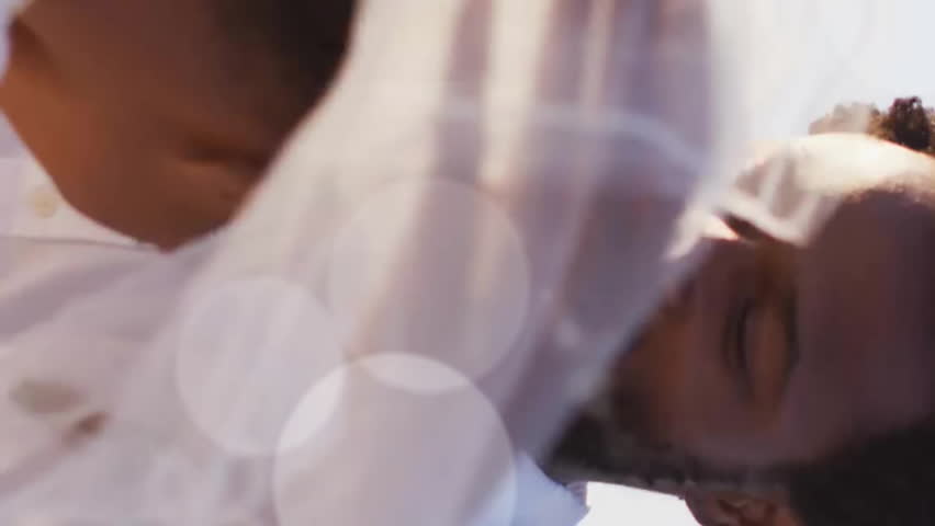 Animation of light spots over happy african american groom unveiling bride on beach at wedding. Romance, love, vacations and marriage, digitally generated video.
