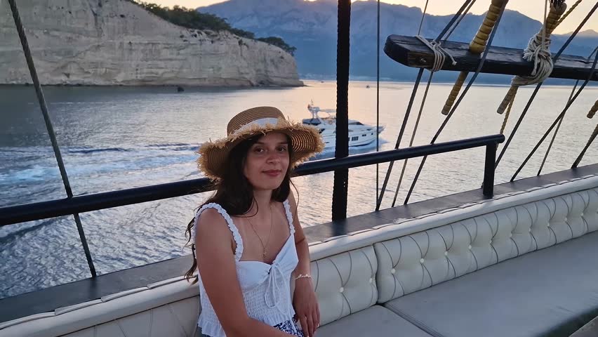 Relaxed young woman tourist wearing a straw hat, sailing in a voyage boat, looking out at the sea and mountains in the background under a sunset sky. Beautiful Turkish Mediterranean coastline of Kemer
