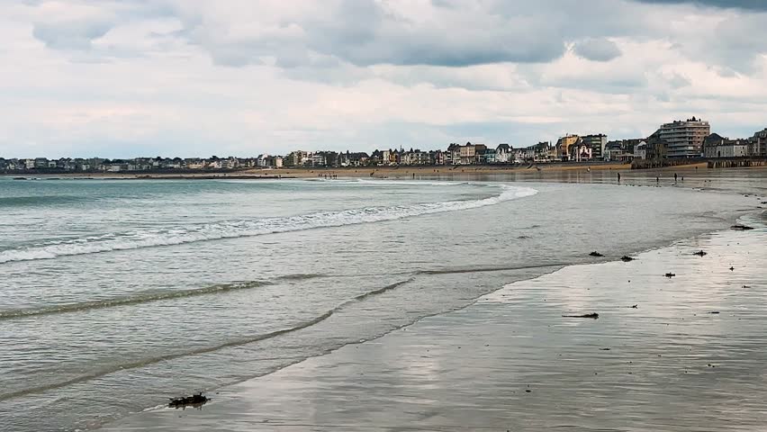 Mirror-like surface of wet sand and small waves at low tide in the English Channel on a Plage de l’Eventail beach overlooking the Intra-Muros district of Saint-Malo, Brittany, France.