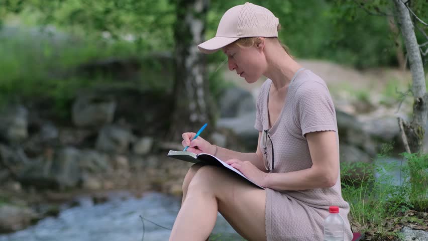 A pensive woman writing in her diary while sitting by water's edge in the forest. A young female hiker writes about her trip. Plans, goals, and memories remain on paper.