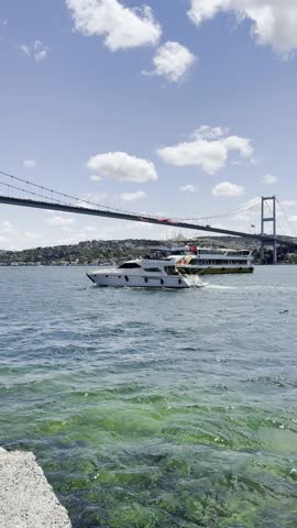 Luxury White Yacht Cruises Gracefully Under 15 July Martyrs Bridge Over Clear Greenish Bosphorus Water While Large Passenger Ferry Maneuvers Alongside Bridge on Bright Sunny Day. Place for Logo, Text.