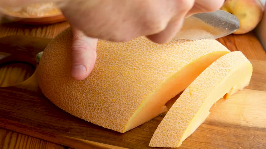 Man cuts ripe yellow melon into pieces on a wooden cutting board using a sharp kitchen knife. Juicy fruit preparation. Seasonal harvest of fresh fruits and berries. Healthy vegetarian food.