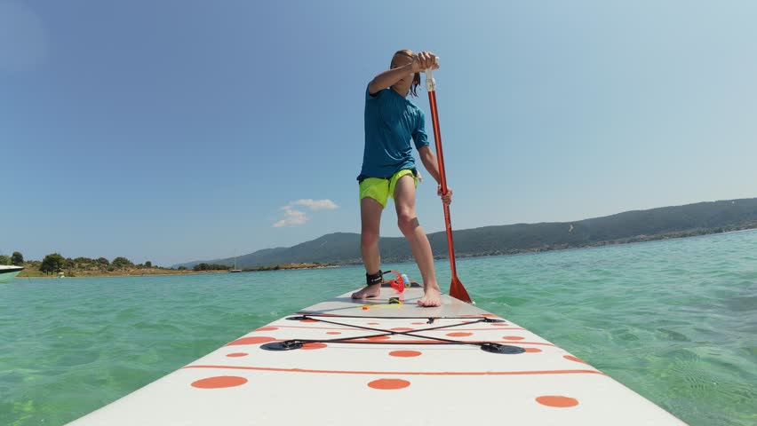 Young boy confidently paddling on a stand-up paddleboard in shallow turquoise sea near a rocky shore. Active summer vacation with water sports and clear sunny skies