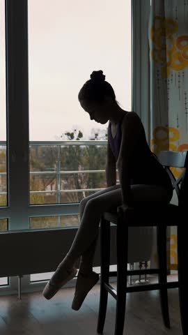 Dark silhouette of Little ballet dancer, girl in a ballet leotard, resting sitting on chair after practicing pointe exercises . ballet lessons at home, online classes. near large window.