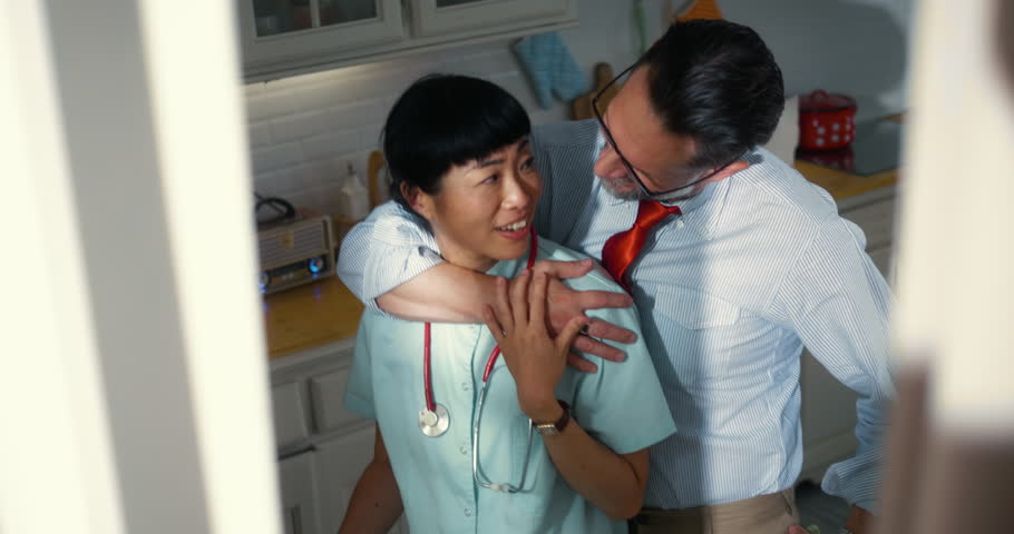 Japanese nurse and her Caucasian businessman husband share a happy moment in the kitchen, laughing and hugging before heading off to work. A joyful multiracial couple enjoying their morning together.
