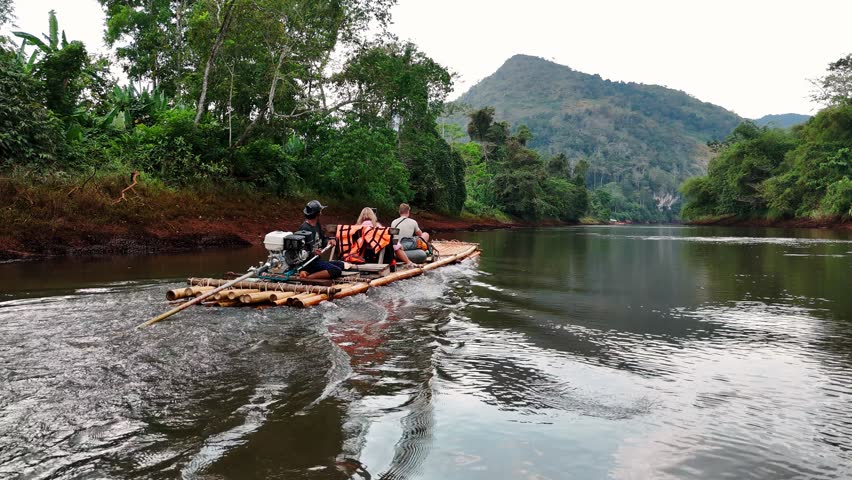 Travelers rafting bamboo raft across serene river, drifting through verdant tropical scenery during sunny day in traditional southeast asian waterway exploration, Thailand