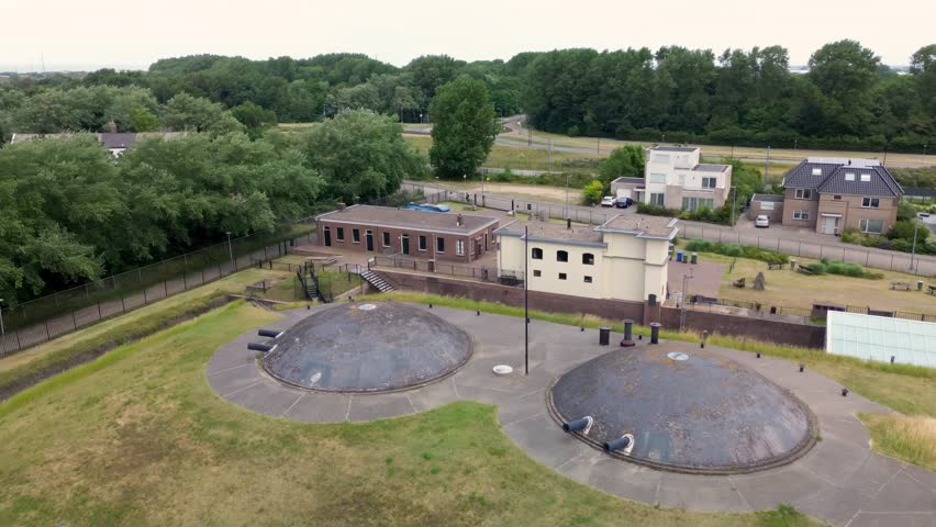 Aerial view of a historic military fort with grass-covered bunkers, surrounded by modern buildings, trees, and walking paths, near the coastline under a bright blue sky.