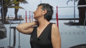 Senior woman with grey hair wearing a sleeveless dress, touching her neck while standing thoughtfully on a seaside promenade with blurred beach and fitness equipment in the background. - Powered by Shutterstock - Get 15% off with code: PIKWIZARD15