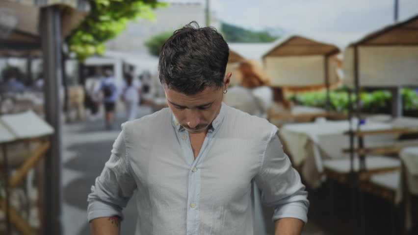 Young hispanic man with a thoughtful expression brushes his hair with his hand while standing on a terrace at an outdoor restaurant, showcasing a relaxed yet stylish vibe.