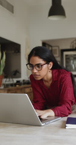 Positive smart young Indian student girl in stylish glasses typing on laptop at home, writing essay, article, turning look away for inspiration, thinking on creative idea. Vertical shot