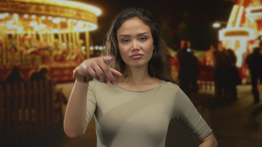 Woman gestures loser sign at night fair with carousel and lights in the background.