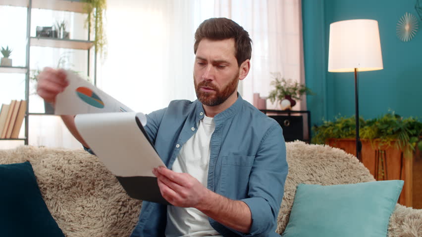 Man at home sits on the sofa, anxiously flipping through bills and documents searching for something crucial. Caucasian guy looks worried trying to find an important paper among financial statements.