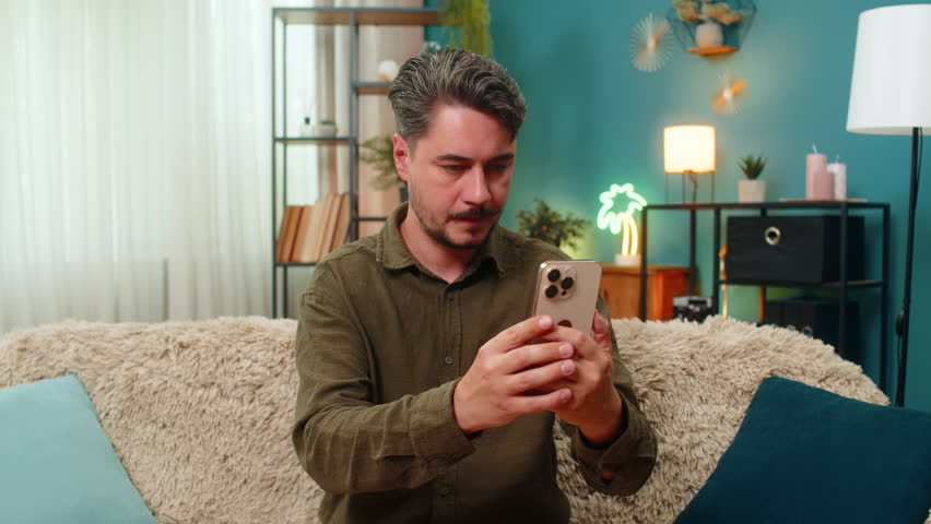 Middle-aged man at home reads smartphone message rolls eyes sighs showing irritation frustration over bad news. Young adult guy on sofa holds mobile phone feeling angry stress tension disappointment