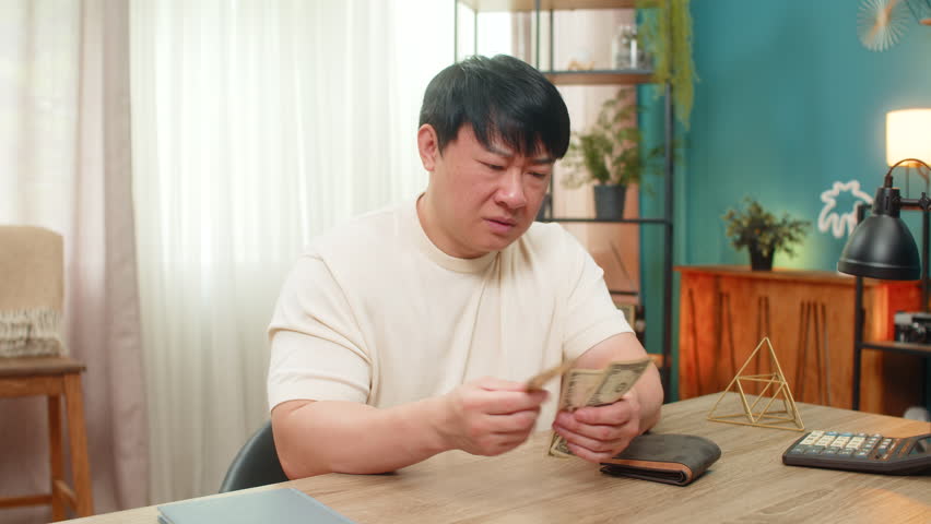 Asian man sitting at home table opens wallet, finds only small amount of money dollar cash, frowns shakes head disappointed. Chinese guy shows ironic discontent, sighs, considering financial shortage