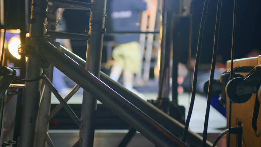 Side close-up of stage metal rig at night with blurred performer in background. No audience visible. Great for backstage, technical, or abstract concert visuals.