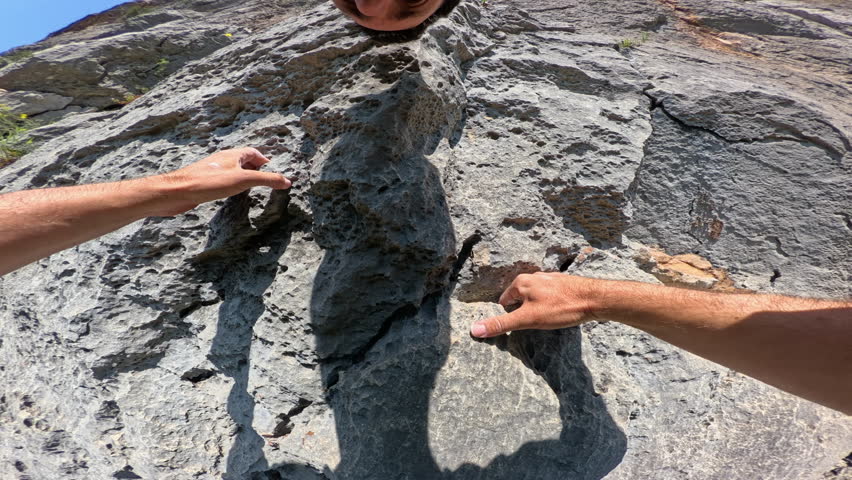POV: Strong male arms climb firmly up a textured rock face, using a quickdraw and blue rope for safety. A look at the technical aspect and equipment used in rock climbing, a challenging outdoor sport.