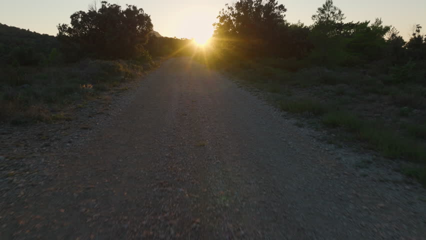 AERIAL: Gravel road winds through forested landscape, directly into warm, golden light of a rising sun. Sunbeams create a beautiful glow, illuminating the path and casting long shadows through trees.