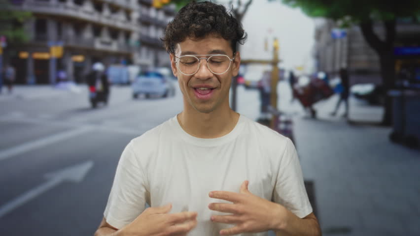 Man wearing glasses with hands on head on street lined with buildings and cars, looking at camera; surprise.