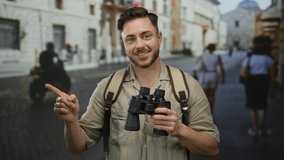 Young man with beard and binoculars smiles on city street pointing while wearing backpack conveying tourist adventure setting on a sunny day outdoors - Powered by Shutterstock - Get 15% off with code: PIKWIZARD15