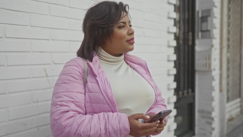 Young woman in pink jacket using smartphone, standing outside against urban brick wall, city street setting, showcasing plus size fashion, casual style outdoors.