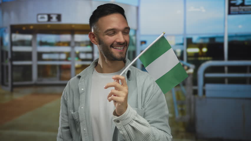 Young man smiling and waving nigerian flag at airport terminal captures a vibrant multicultural travel experience.
