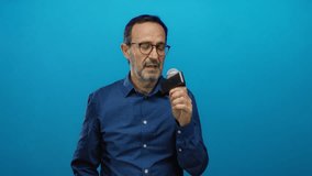 Middle-aged man in blue shirt holding microphone against isolated blue background presents confidently. - Powered by Shutterstock - Get 15% off with code: PIKWIZARD15