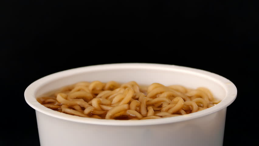Cooked noodles lifted by chopsticks from glossy beige cup on black background in tight close-up, for instant food packaging.