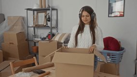 Woman unpacking boxes in new home with headphones, enjoying music, surrounded by cardboard boxes, furniture, and books in cozy living room setting. - Powered by Shutterstock - Get 15% off with code: PIKWIZARD15