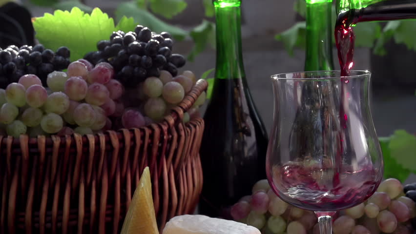 Red Wine Poured in a Glass. In pouring a glass of wine on the background of a basket with grapes, wine bottles and pieces of cheese. Slow Motion