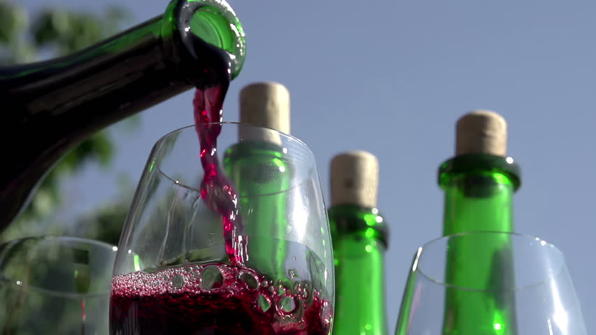 Pouring Red Wine in Goblet. Red wine pouring down from a bottle into a glass on a background of wine bottles. Slow Motion