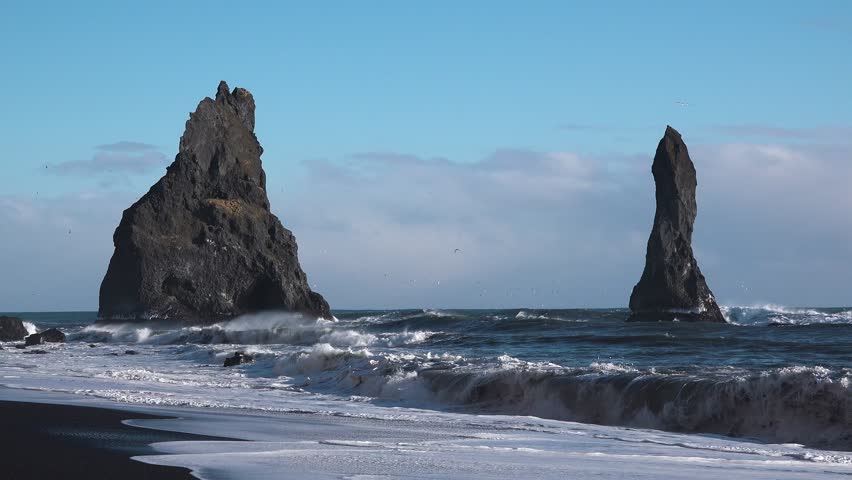 Roiling waves crash against black sand as towering rock formations rise from the ocean on a stormy day. The dramatic coastal scene showcases nature