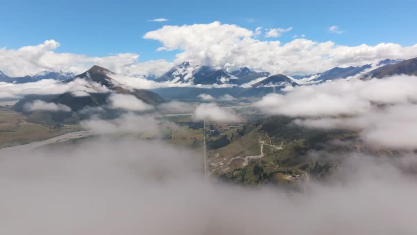 An awe-inspiring aerial perspective of a mountainous region partially covered by thick clouds and fog, revealing green hills, winding roads, rocky and snow-capped peaks, and a river meandering through
