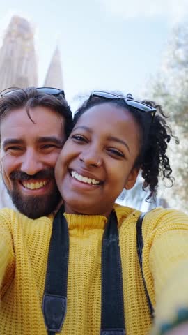 Multiracial young tourist couple taking a selfie vertical video in front of the sagrada familia, enjoying their vacation together, looking at camera smiling. Copy space.