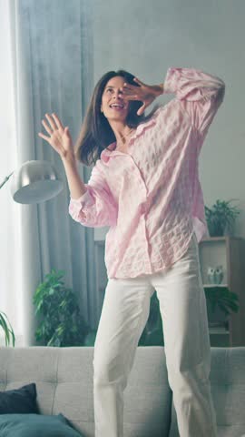 Young woman dances energetically in a bright, modern living room with vibrant green plants and stylish decor, creating a lively and uplifting atmosphere. 4K video, slow motion, 50 fps