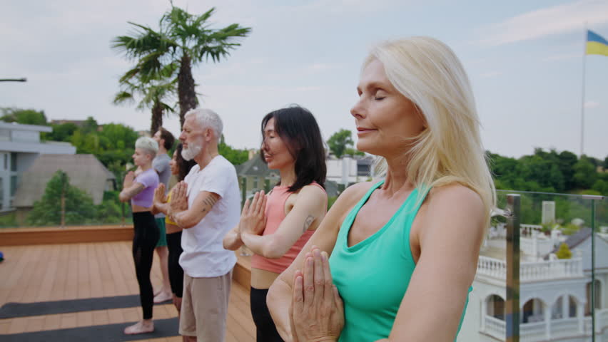 Smiling blonde mature woman practices yoga with multiracial group on large hotel terrace. Different aged sportive people train body and mind against city