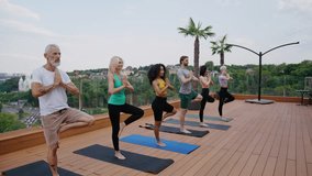 Trained multicultural sportspeople stand in yoga tree positions on hotel rooftop. Different aged friends practice body and mind care against city park - Powered by Shutterstock - Get 15% off with code: PIKWIZARD15