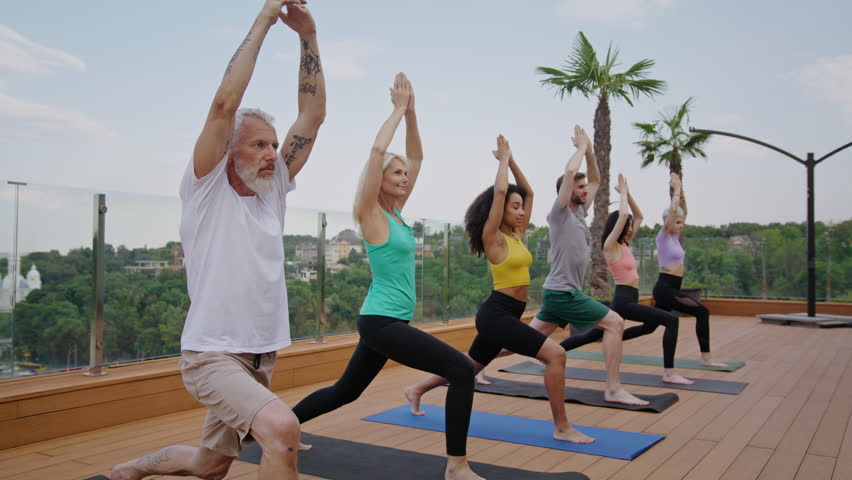 Diverse people in activewear stand in warrior poses on hotel rooftop terrace. Different aged yoga practitioners exercise with mindfulness against urban park - Powered by Shutterstock - Get 15% off with code: PIKWIZARD15