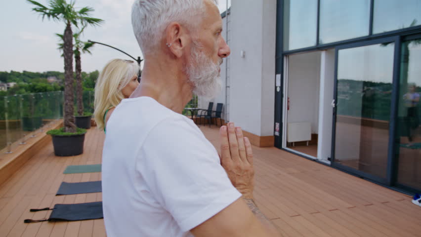 Old bearded man stands in yoga tree asana with multiethnic group on hotel terrace. Elderly male athlete practices mindfulness with friends at exotic resort