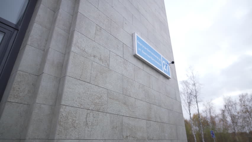 Building wall features address sign with street name and number on tile surface. Camera tilts upward capturing blue signage against sky