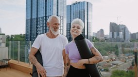 Senior man and young woman stand on rooftop smiling and holding yoga mats after fitness session. Modern city buildings provide location of outdoor yoga activity - Powered by Shutterstock - Get 15% off with code: PIKWIZARD15