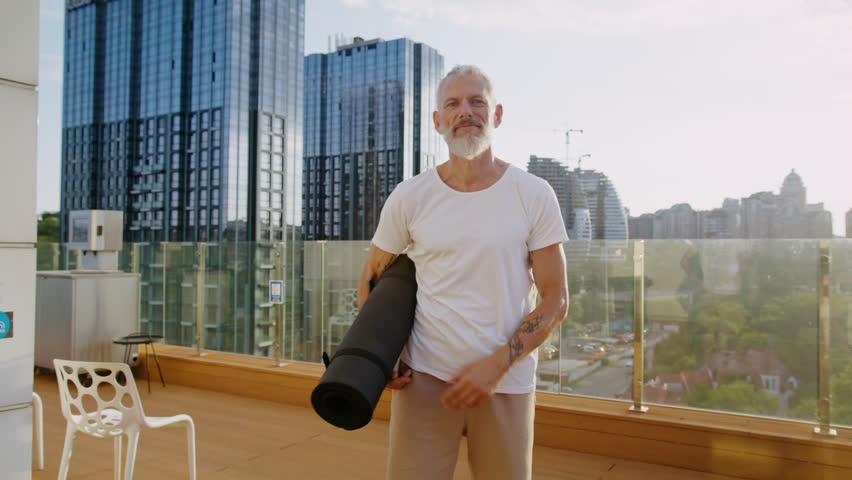 Happy senior man stands on rooftop with city buildings on background holding rolled yoga mat. Looking at camera smiling suggests photoshoot after workout - Powered by Shutterstock - Get 15% off with code: PIKWIZARD15