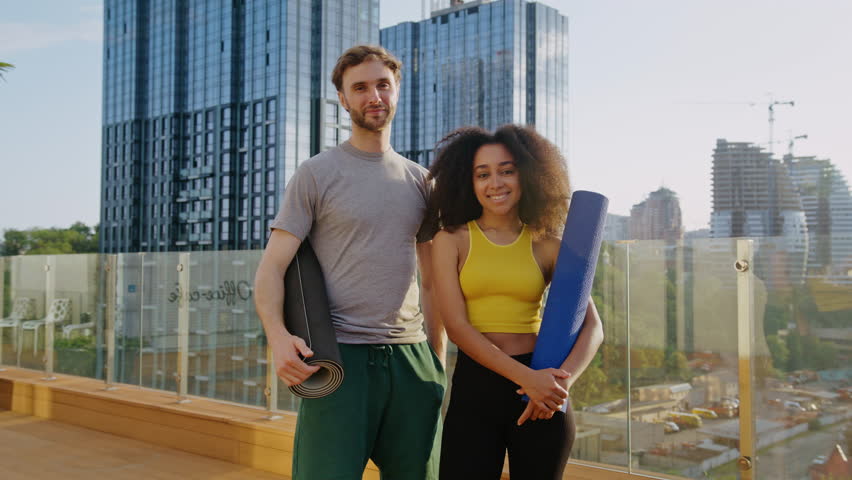 Bearded man and African American woman stand side-by-side on rooftop holding yoga mats by city. Warm smiles suggest friendly and shared interest in wellness - Powered by Shutterstock - Get 15% off with code: PIKWIZARD15