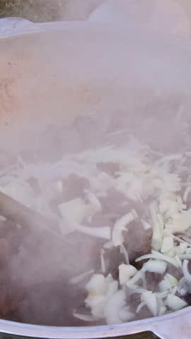 Close-up of a large pot with meat and chopped onions being stirred, generating significant steam while cooking outdoors on grass with a gas canister nearby. Hand using a perforated spatula to mix