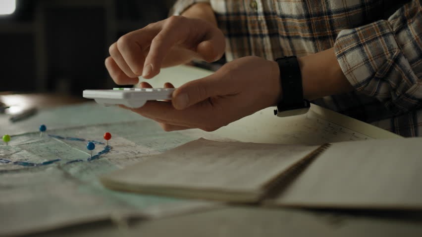 A person focuses on calculating figures using a calculator while surrounded by maps and notes in a low-light setting, indicating careful planning or analysis