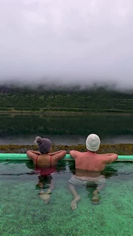 A couple of men and women soak in a beautiful geothermal hot spring, surrounded by lush green hills and misty mountains in Iceland. Hot pool, Westfjords, Iceland