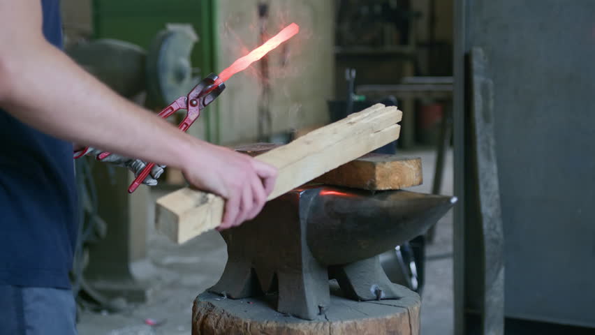 Side angle of blacksmith hammering hot metal with wood on anvil. Captures traditional forging technique, heat, sparks, and craftsmanship in an authentic workshop setting.