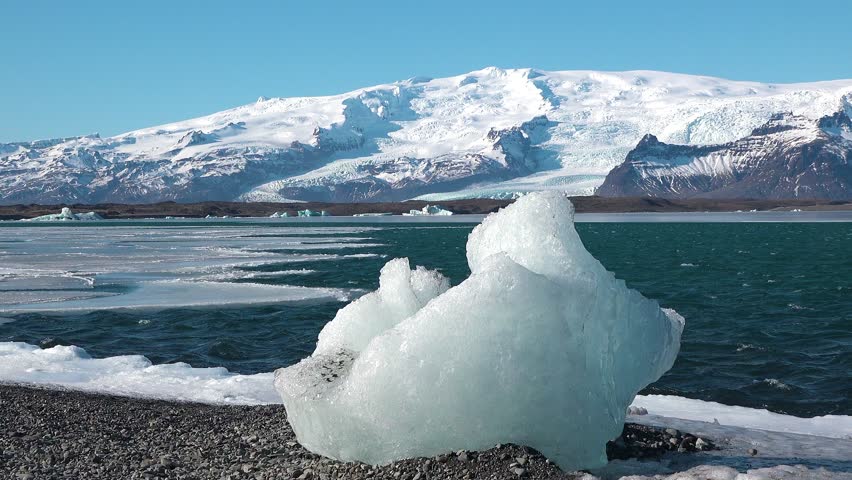 A stunning landscape features a contrast of icy formations along the shoreline and vibrant blue waters with majestic snow-covered mountains in the background.